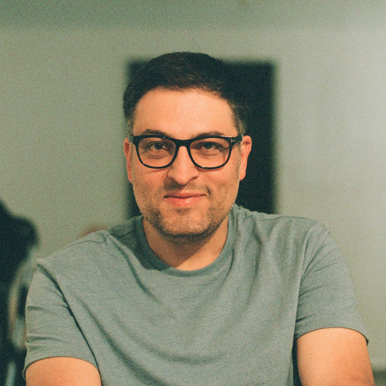 Man wearing glasses and a grey t-shirt smiling while sitting indoors.
